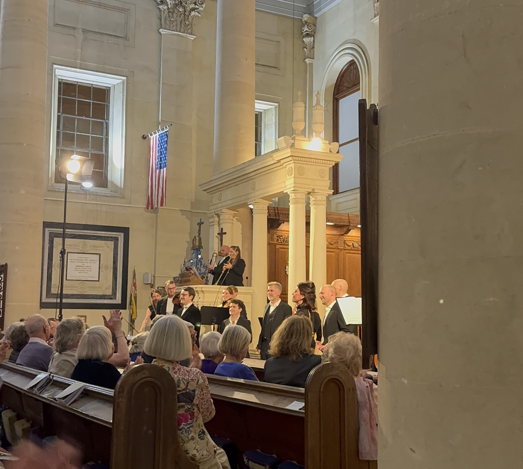 Choir performing in a church nave beside ornate columns and an illuminated organ loft, with seated audience out front.