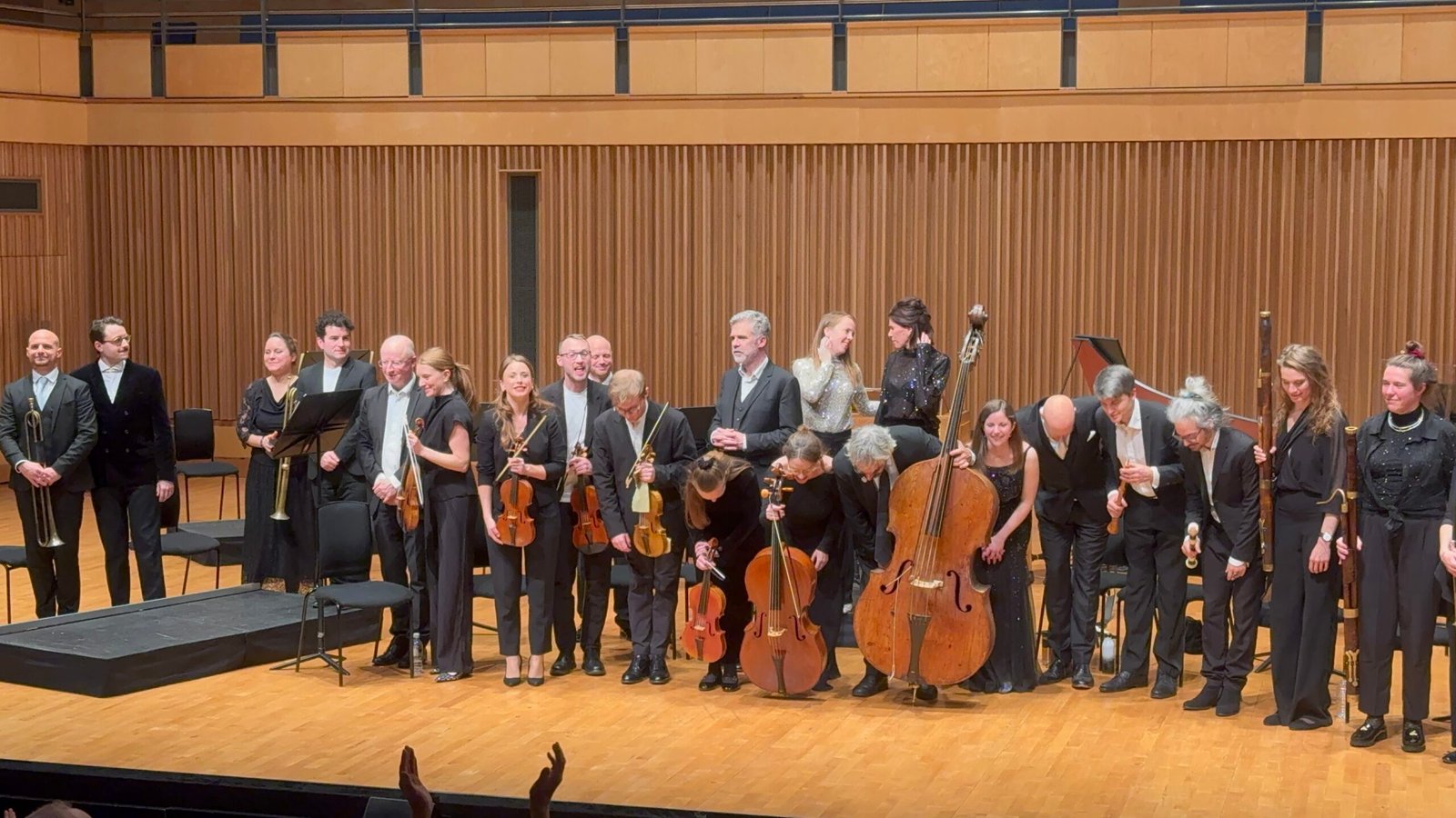 Orchestra group in formal black on a wood stage after a concert, bowing and smiling with string instruments like violins and cellos visible.