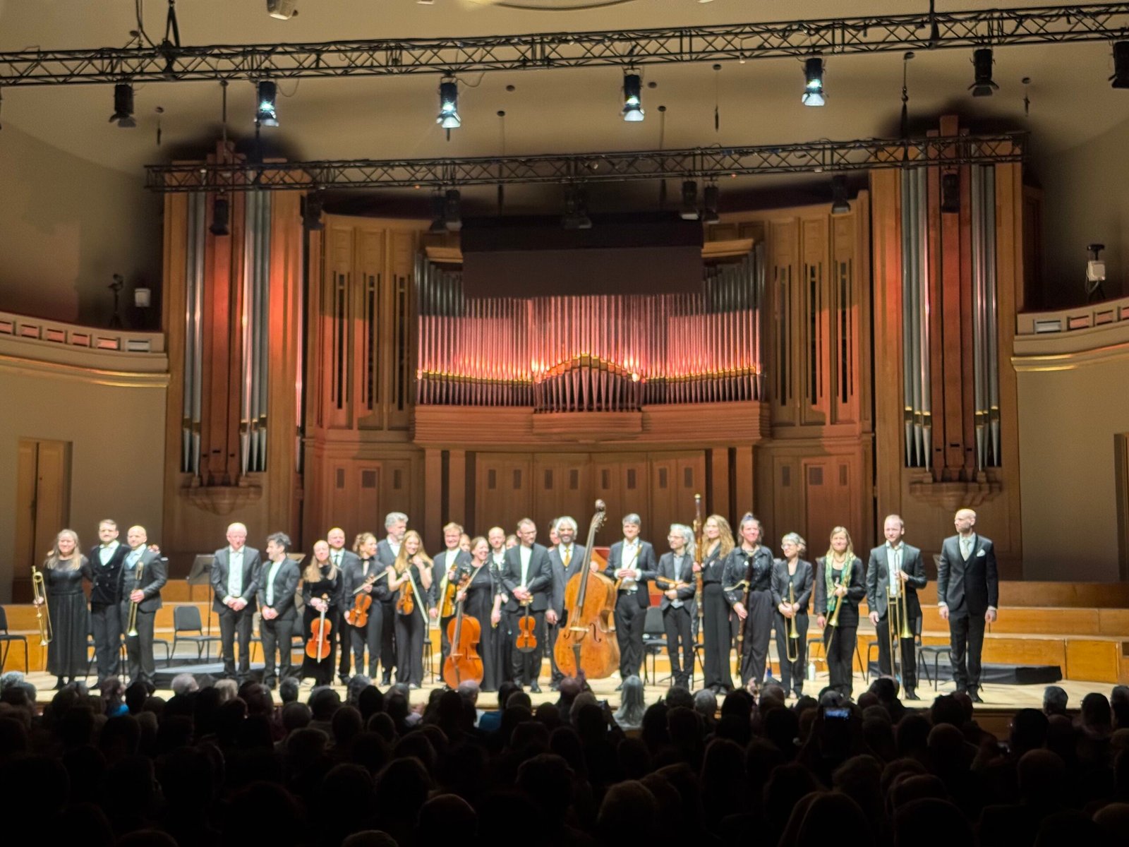 Large choir and string orchestra on stage in a concert hall, audience visible in the foreground.