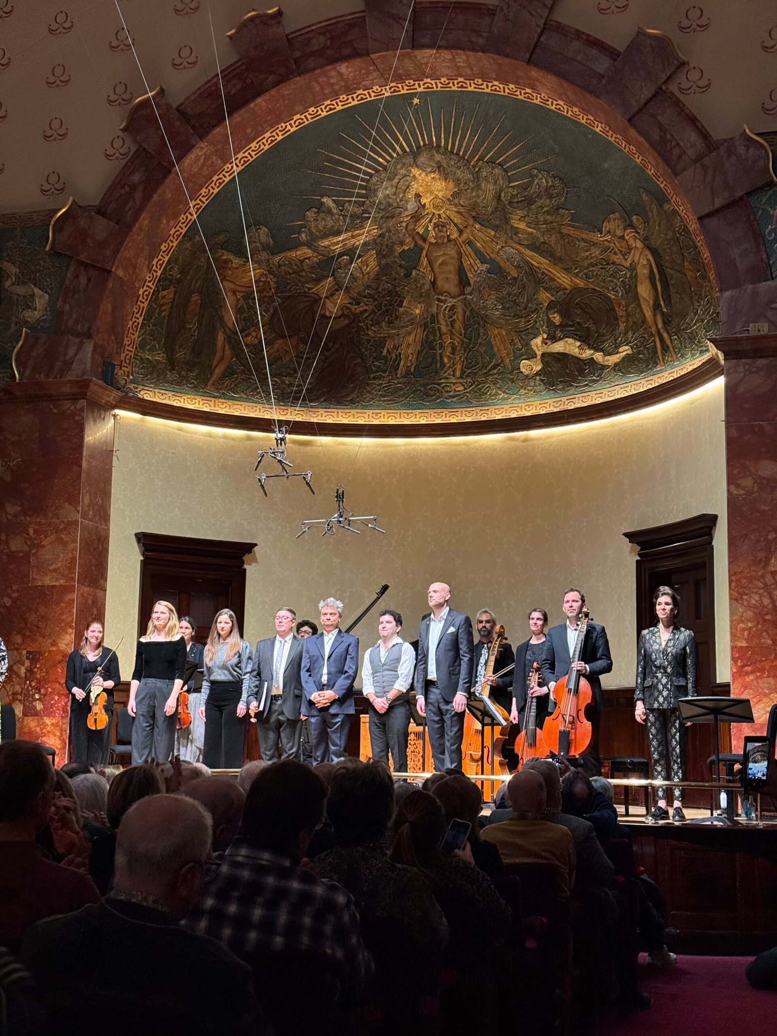 Concert hall stage with string musicians standing, holding violins and a cello, audience in foreground, ornate semicircular mural above them