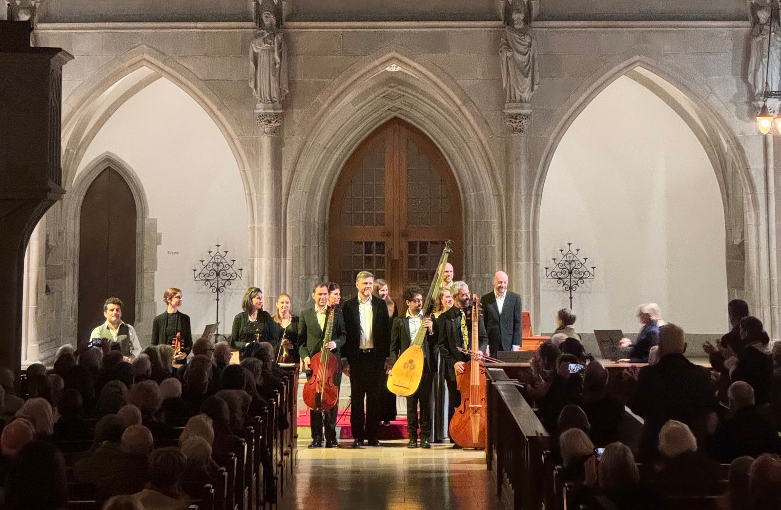 Choir and musicians stand on a church stage with stone arches, instruments in hand, facing a seated audience.
