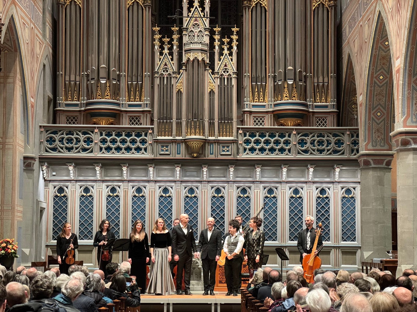 Vocal ensemble in formal dress standing on a decorated stage with a Gothic organ backdrop and an audience watching.",