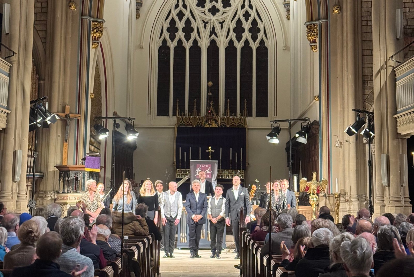 Choir performs in a Gothic church with an ornate altar and stained arches, facing a seated audience.