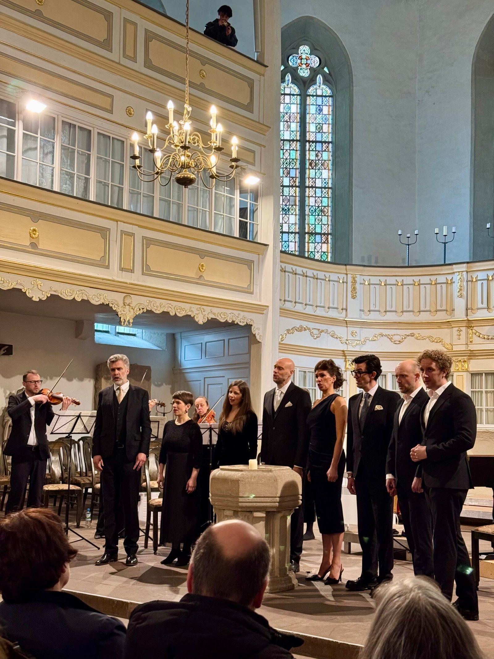 Choir in formal black attire sings in a historic church with gold-trimmed balconies and a large chandelier above, audience seated nearby.