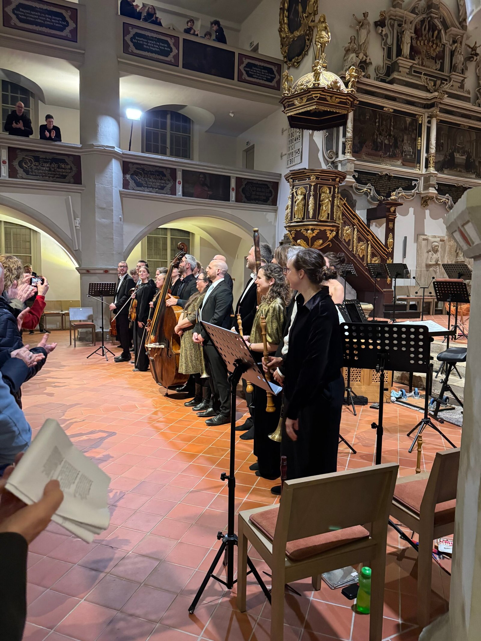 A formal choir and musicians stand at the front of a church nave with ornate gold pulpit and baroque wall carvings; audience claps on the left and a person in the foreground shoots a photo.