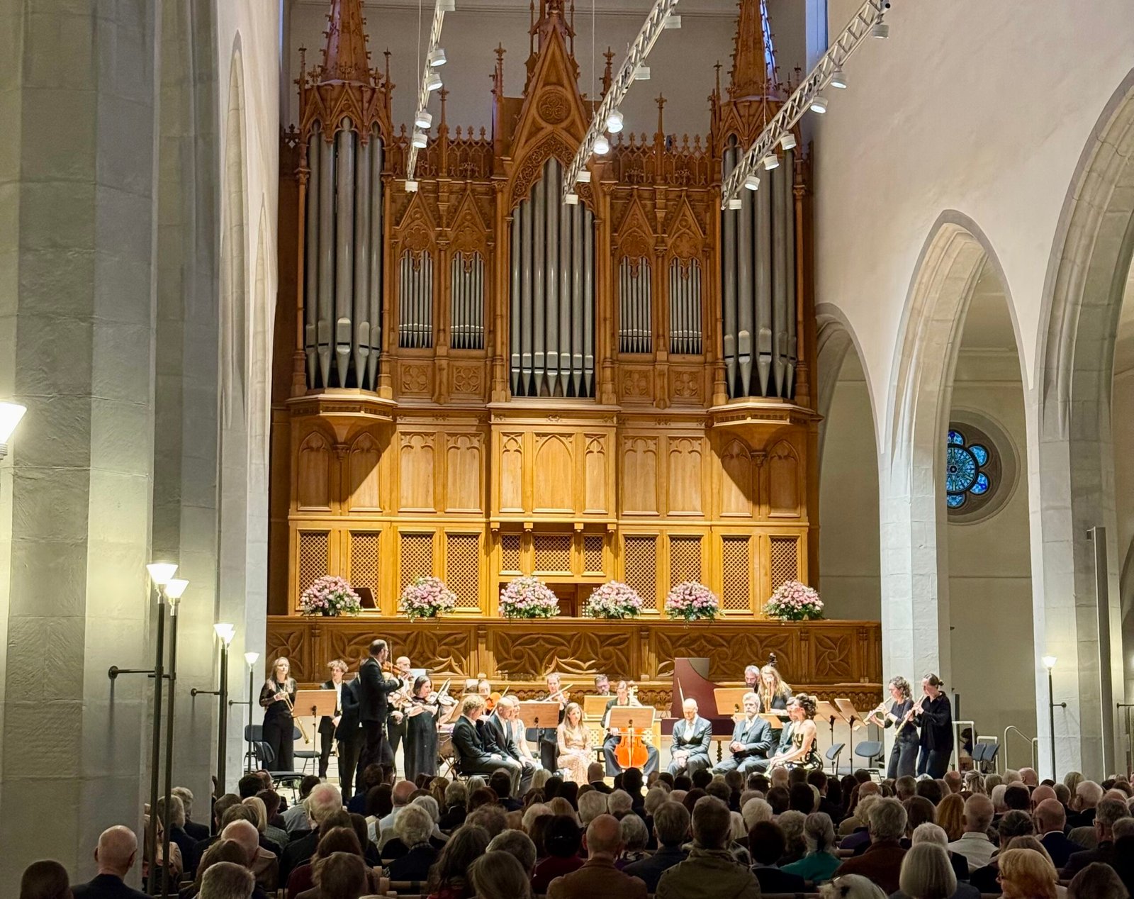 Orchestra performing in a church with a large ornate wooden organ behind them and a seated audience.