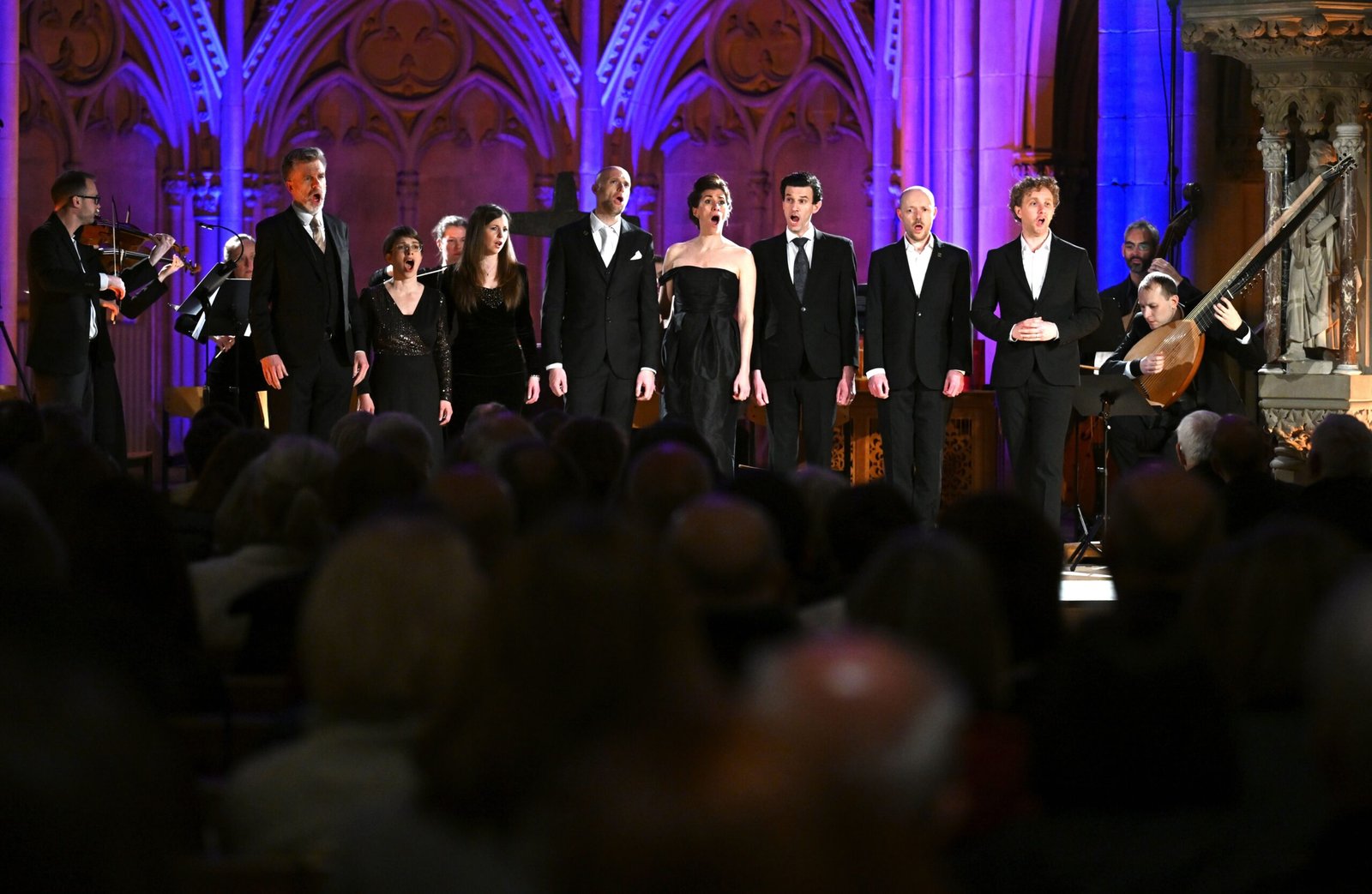 Choir in black formal wear performing on a lit stage inside a church-like venue with purple-blue lighting and audience in the foreground.