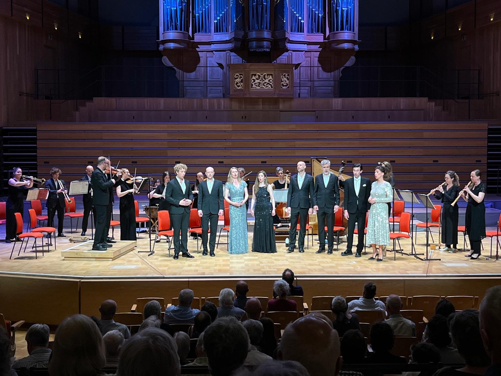 Orchestra members in formal attire stand on a stage in a concert hall, with an organ backdrop and empty red chairs behind them.