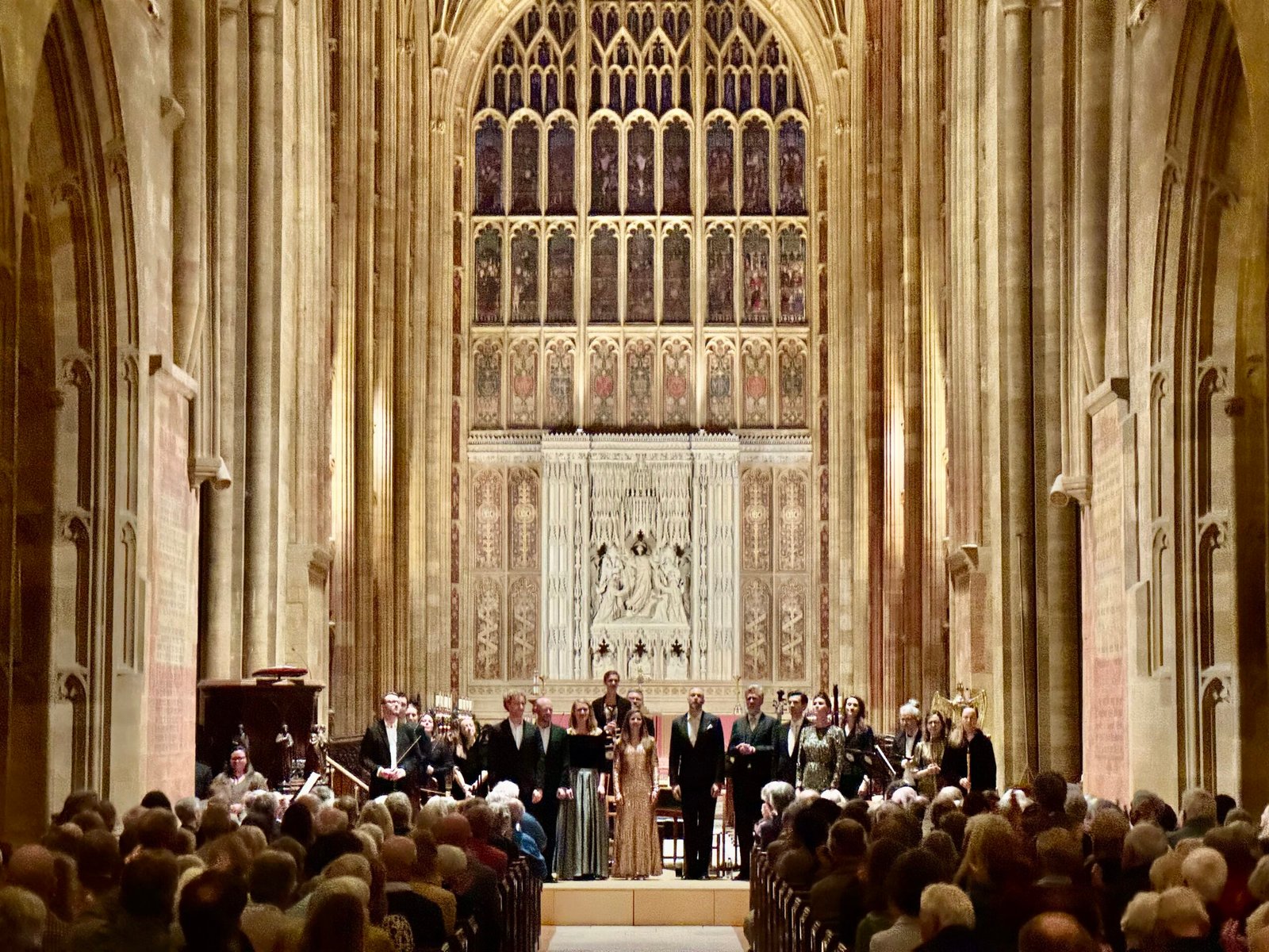 Chorus of formally dressed singers performing on a cathedral stage, with ornate stone arches and a filled audience in the foreground.