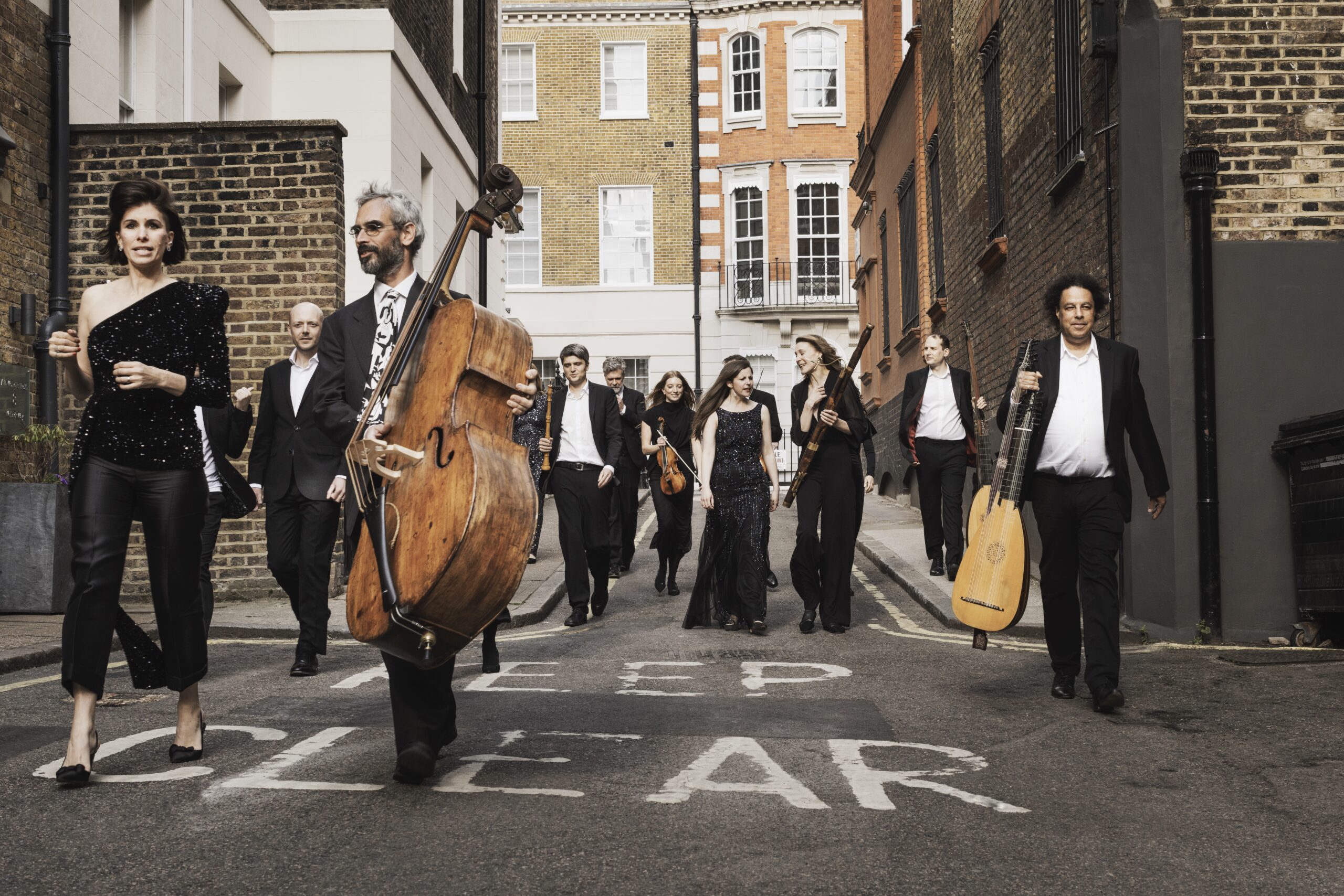 A group of formally dressed musicians walking down a narrow brick alley, carrying a double bass and various string instruments.