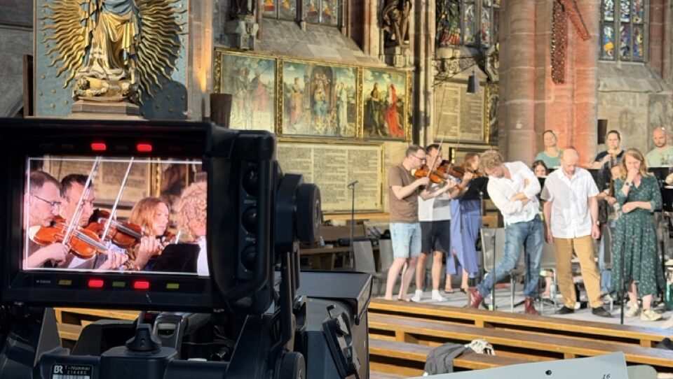 Video camera records a live choir and violinists performing in a church with colorful stained glass windows behind them? no